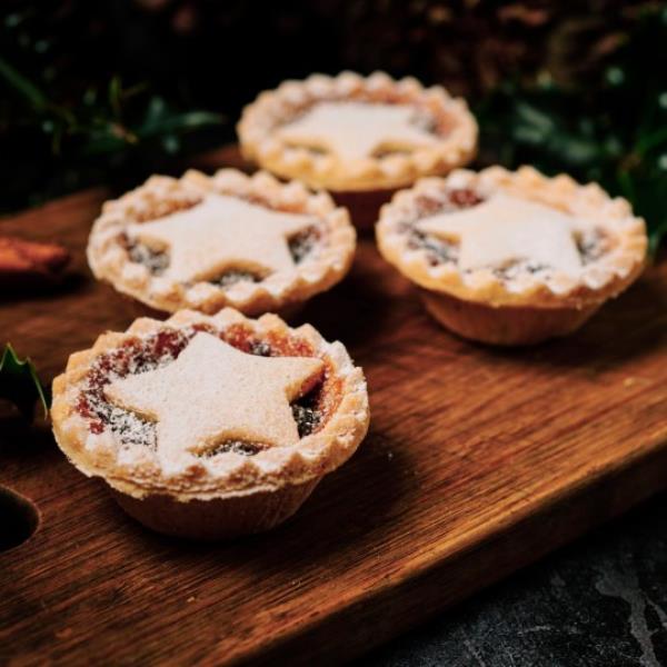 Four Mince pies dusted with icing sugar on a dark wood board with Holly and Pine cones.