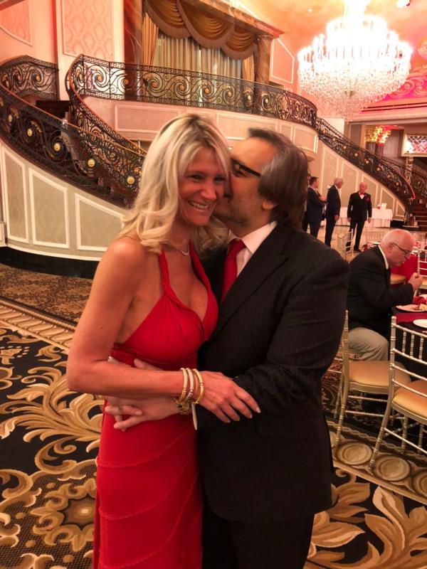 A man appearing to kiss the side of Nadine Arslanian's hair. She is wearing a low-cut red dress, they are standing in front of a staircase in what appears to be the lobby of a theater.