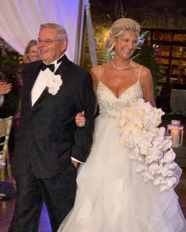 Nadine Arslanian smiling in a low cut strappy wedding dress and holding an arrangement of flowers. She is arm in arm with Bob Menendez who is wearing a tux and prominent buttonhole