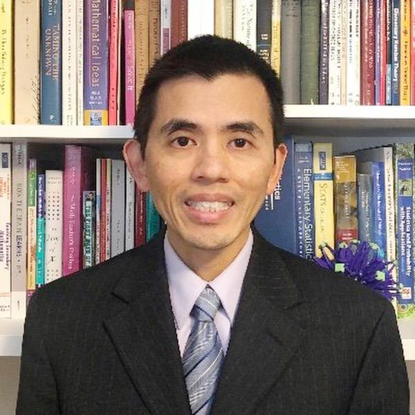 Bobson wong in black suit, white shirt and blue grey tie, smiling and looking at a camera while sitting in front of rows of books on white shelves