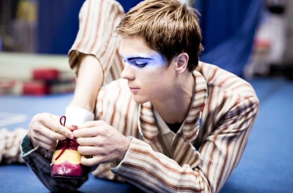 A male co<em></em>ntortionist lying down face first, and twisting his leg around so he can tie his shoe.