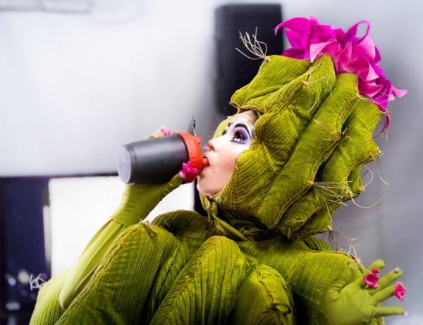 A Cirque performer dressed as a cactus takes a sip from a mug.