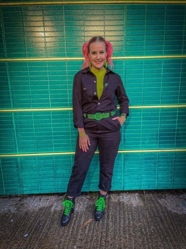 Zara Canfield standing in front of a green brick wall with thin yellow horizo<em></em>ntal stripes; wearing a pantsuit with a bright green belt and a mustard top underneath, left hand in her pocket and right hand by her side, smiling at the camera.