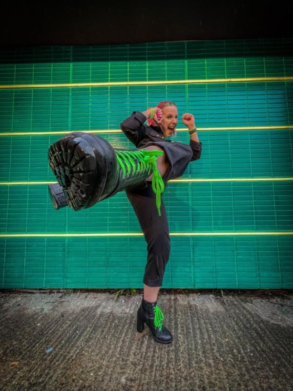 Zara Canfield in front of a green brick wall with thin yellow horizo<em></em>ntal stripes; she's wearing a navy pantsuit and high-heeled black boots with bright green laces, kicking one foot at the camera, both arms raised by her face, hands in fists and smiling at the camera.