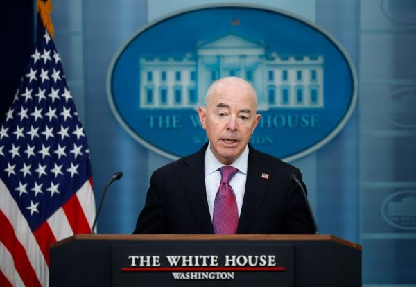 Alejandro Mayorkas speaking at a podium with micropho<em></em>nes and a flag, regarding an assassination attempt on Do<em></em>nald Trump, present.
