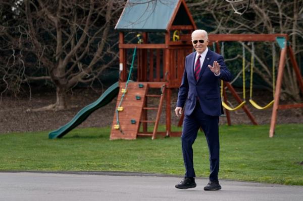 U.S. President Joe Biden in a suit and black HOKA sneakers, waving while walking towards Marine One to depart the White House.