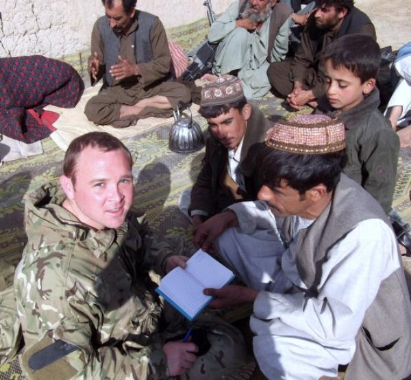 Jo<em></em>nny in 2011 helping train members of the Afghan Natio<em></em>nal Police, sitting on the floor, holding a notebook, with Afghan men and children around him